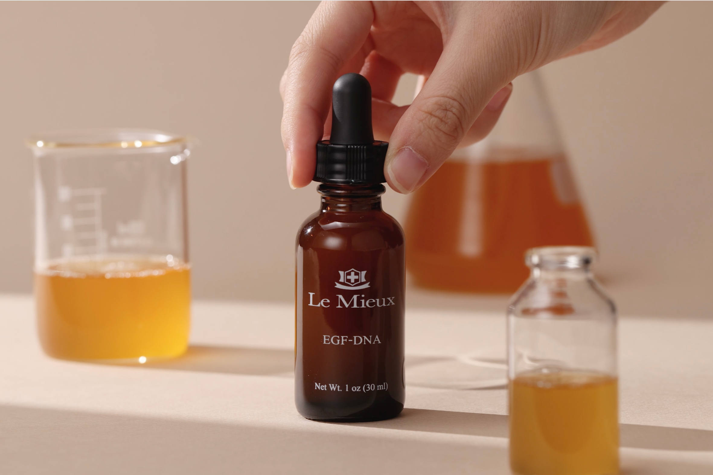 Hand holding a brown bottle labeled 'Le Mieux' with a dropper, next to a glass container and another bottle on a neutral background.