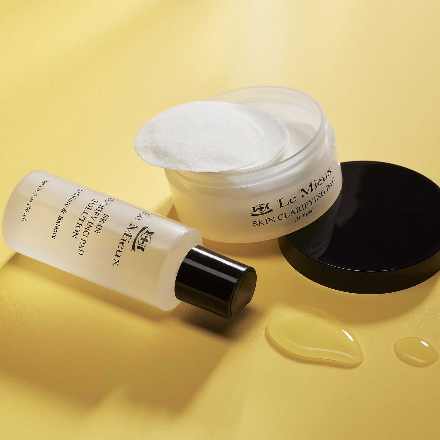 Skin Clarifying Pad Solution (2 oz) resting on it's back behind some drops of solution. Next to them is an open Skin Clarifying Pad container that's tilted against the black lid. All on a yellow backdrop.