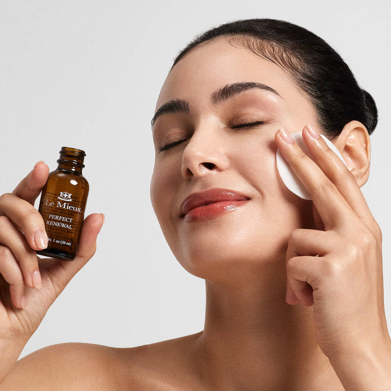 Woman holding a 1 oz Perfect Renewal in one hand and pressing a white cotton pad to her cheek with the other hand.