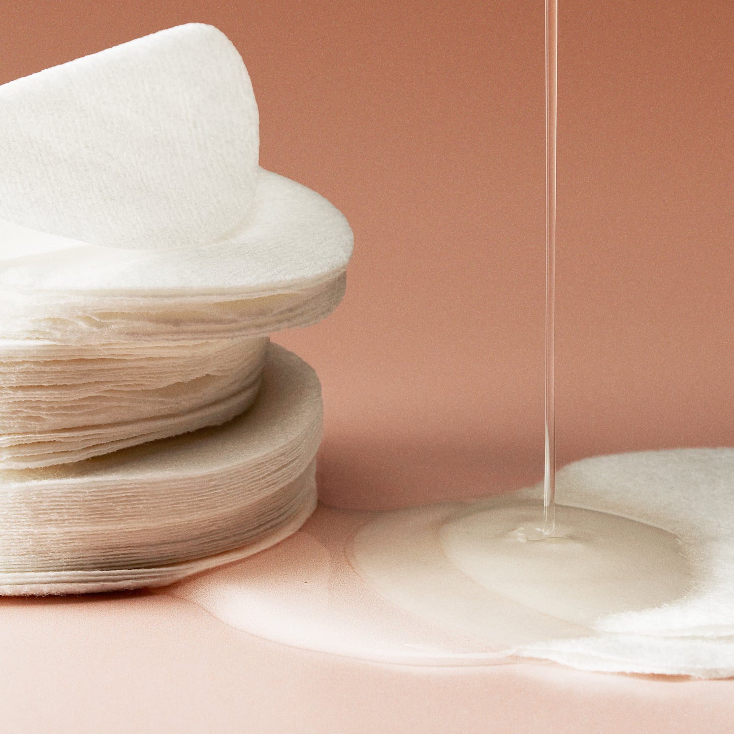 Stack of cotton pads next to two cotton pads with serum being poured onto them in front of a pink background.