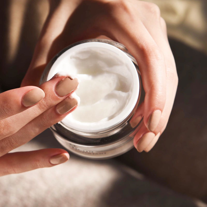 Hand holding an open Microderm Exfoliant while other hand has scooped some of the white exfoliant texture onto fingers.