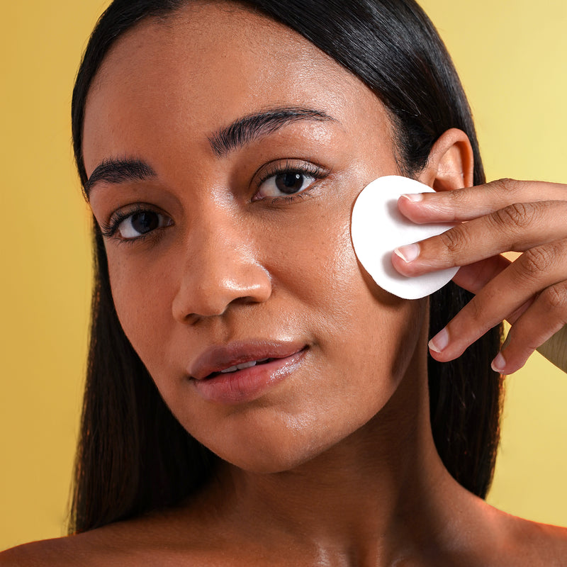 Woman in front of a yellow background with white cotton pad swiping against her cheek.