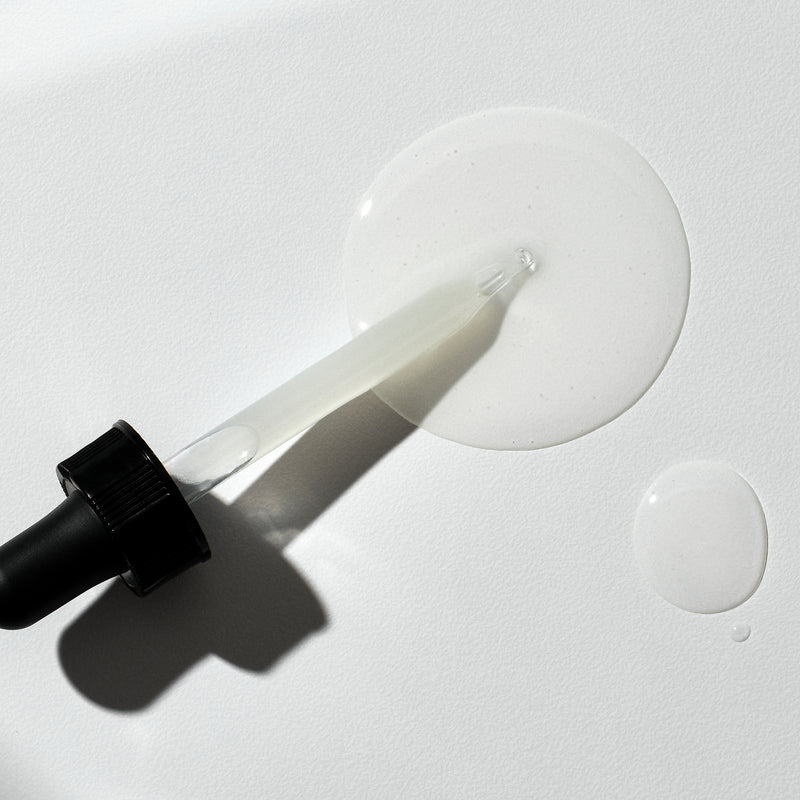 Dropper with black lid and filled with TGF Booster serum laying on a pool of serum on a white background.